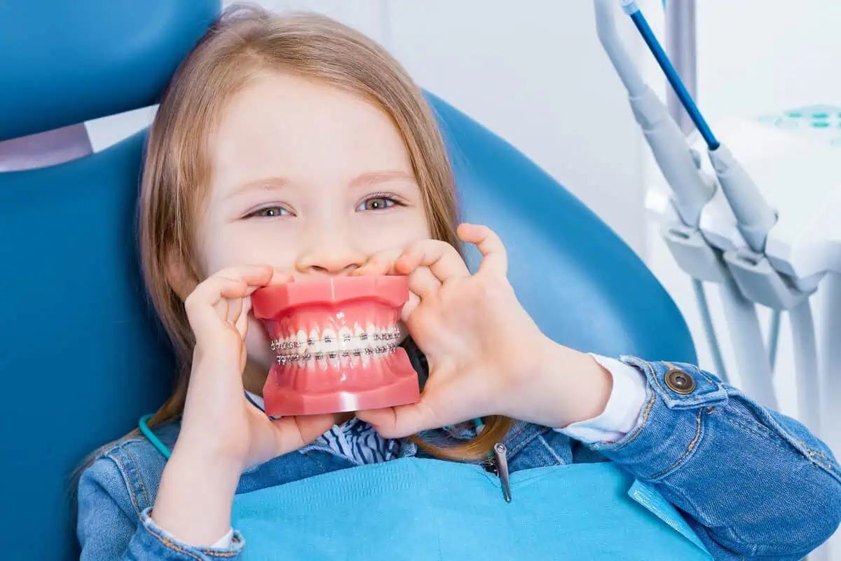 Smiling little girl in the dental chair holding a model of teeth in front of her mouth